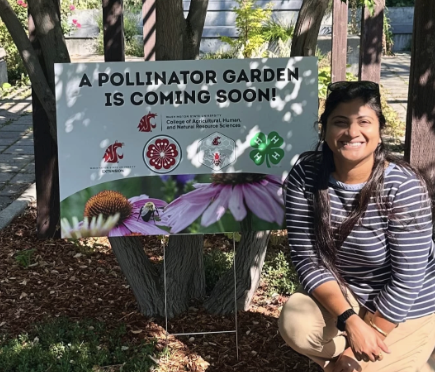 Priya near a "pollinator garden coming soon" sign in the Horticulture Garden by Ensminger Pavilion in Pullman